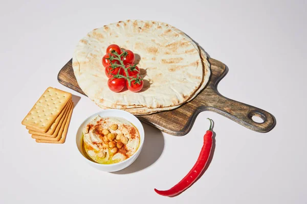 Bowl with hummus, crackers, chili and cutting board with pita bread and cherry tomatoes on grey — Stock Photo