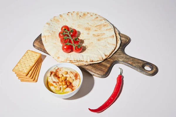 Cuenco con hummus, galletas saladas, chile y tabla de cortar con pan de pita y tomates cherry en gris — Stock Photo