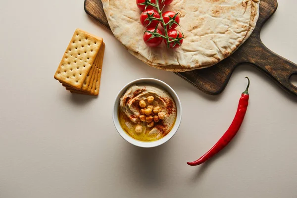 Top view of bowl with hummus, crackers, chili and cutting board with pita bread and cherry tomatoes on grey — Stock Photo