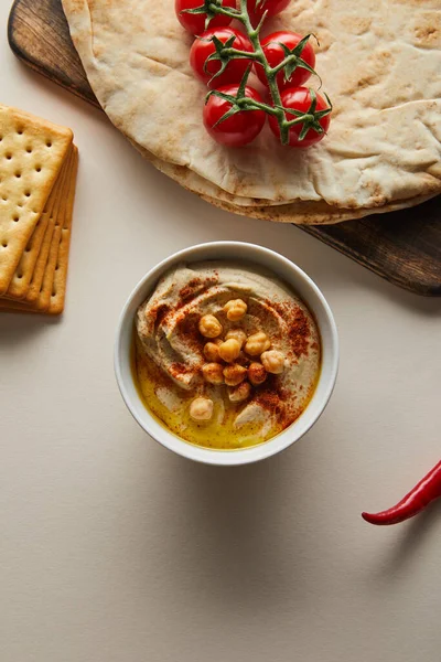 Close up view of bowl with hummus near crackers, chili, pita bread and cherry tomatoes on grey — Stock Photo