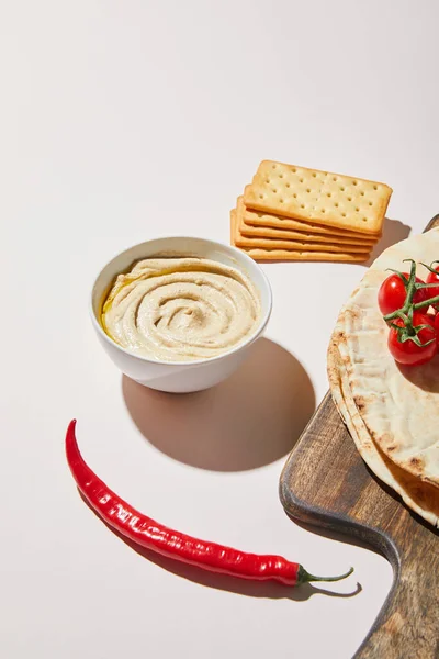 Ciotola con hummus vicino cracker, peperoncino, pane pita e pomodorini su grigio — Foto stock