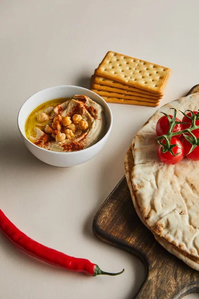 Чаша с хумусом, крекеры, пита хлеб и овощи на разделочной доске на сером фоне — стоковое фото