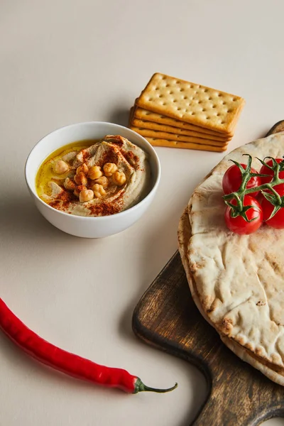 Bowl with hummus, crackers, pita bread and vegetables on cutting board on grey background — Stock Photo