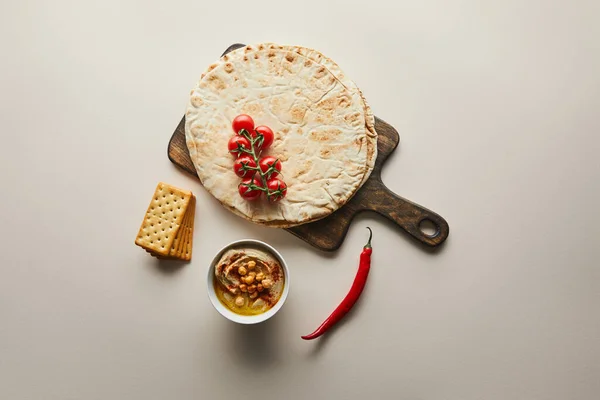 Вид зверху миски з гумусом, крекерами, обробна дошка з хлібом піта та овочами на сірому фоні — стокове фото