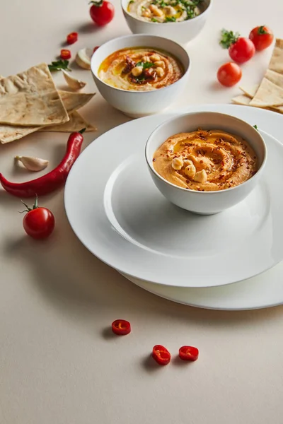 Selective focus of plates, bowls with hummus, cherry tomatoes, chili peppers, garlic and pita bread on grey — Stock Photo