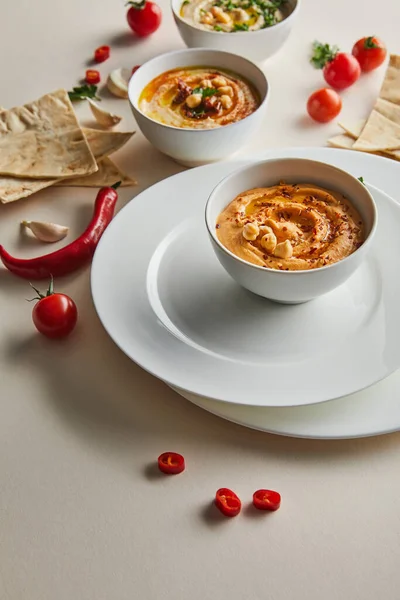 Selektiver Schwerpunkt von Tellern, Schalen mit Humus, Kirschtomaten, Chilischoten, Knoblauch und Fladenbrot auf grau — Stockfoto