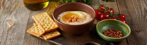 Crackers, bowls with hummus and spices on cutting board, cherry tomatoes, jar of olive oil on wooden background, panoramic shot — Stock Photo