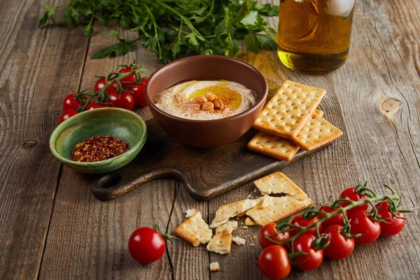 Crackers, ciotole con hummus e spezie su tagliere, verdure e vasetto di olio d'oliva su fondo di legno — Foto stock