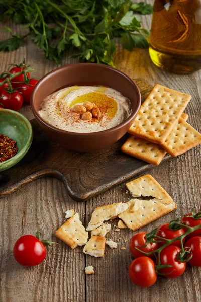 Cracker, Schalen mit Humus und Gewürzen auf Schneidebrett, Gemüse, Petersilie und einem Glas Olivenöl auf Holzgrund — Stockfoto