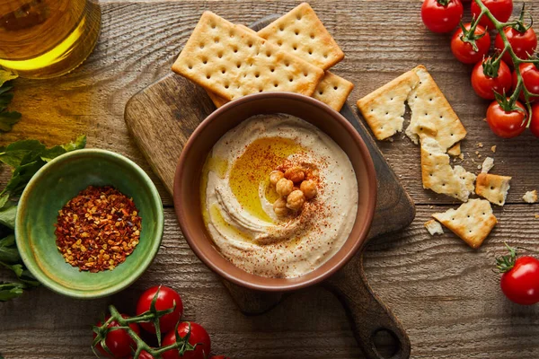 Top view of crackers, bowls with hummus and spices, vegetables, parsley and jar of olive oil on wooden background — Stock Photo