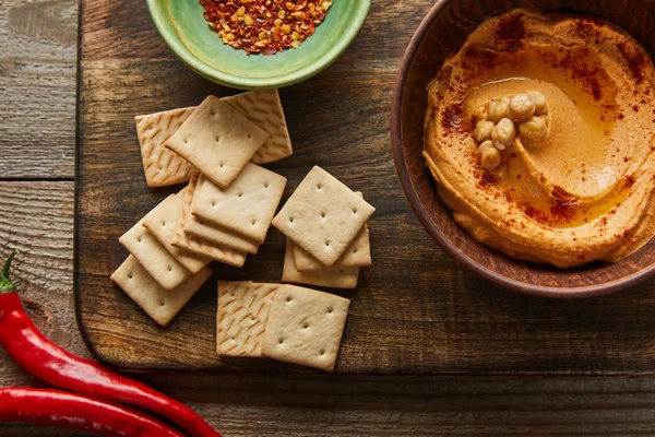 Top view of crackers, bowls with paprika and hummus on cutting board with chili on wooden background — Stock Photo