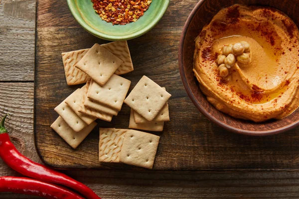 Vue de dessus des craquelins, bols avec paprika et houmous sur planche à découper avec chili sur fond en bois — Photo de stock