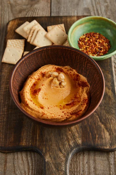 Selective focus of crackers, bowls with paprika and hummus on cutting board on wooden background — Stock Photo