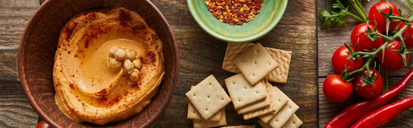 Top view of crackers, bowls with spices and hummus on cutting board with vegetables on wooden background, panoramic shot — Stock Photo