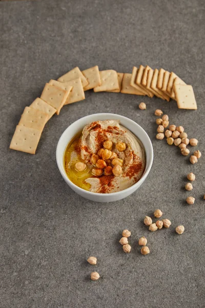 Tigela com hummus saboroso, grão de bico e biscoitos no cinza — Fotografia de Stock