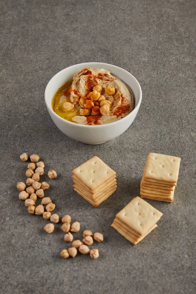 Bowl with delicious hummus near chickpea and crackers on grey — Stock Photo