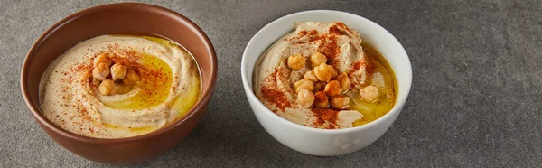 Bowls with tasty hummus with chickpea, spices and olive oil on grey, panoramic shot — Stock Photo