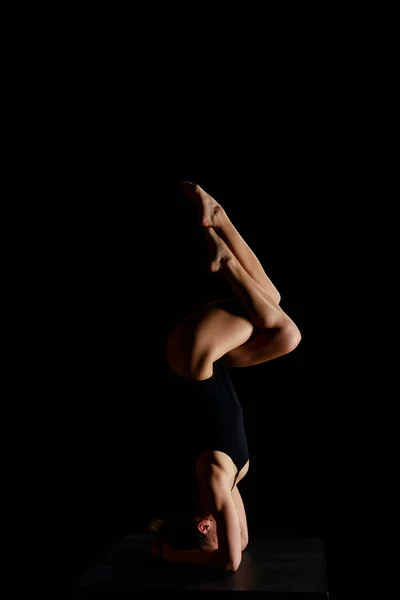 Girl in bodysuit doing eagle legs handstand exercise isolated on black — Stock Photo