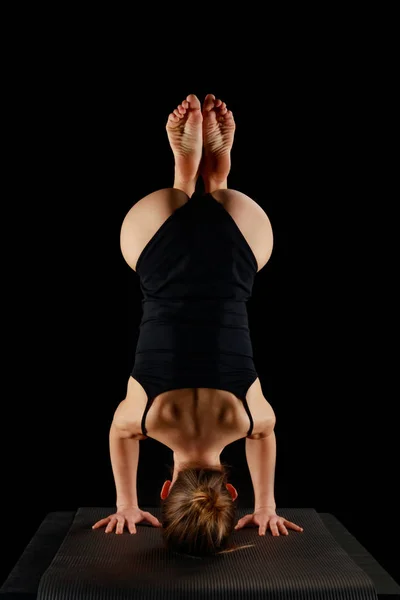 Back view of girl in bodysuit doing yoga headstand exercise isolated on black — Stock Photo