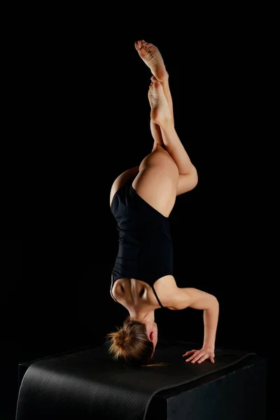 Girl in bodysuit doing eagle legs handstand exercise isolated on black — Stock Photo