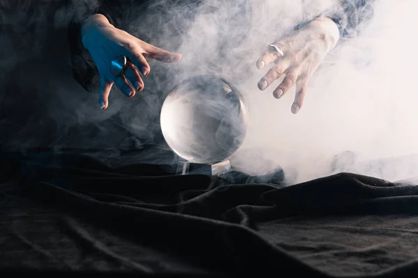 Vista recortada de bruja realizando ritual con bola de cristal sobre fondo oscuro - foto de stock