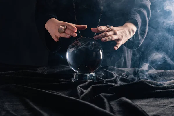 Vista cortada de bruxa realizando ritual com bola de cristal no fundo preto — Fotografia de Stock