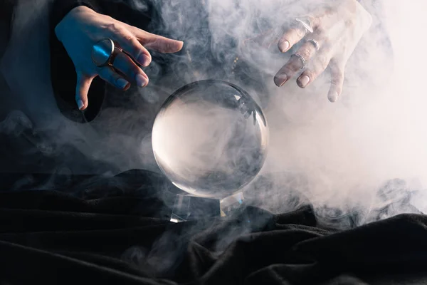 Cropped view of female hands above crystal ball with smoke on dark — Stock Photo