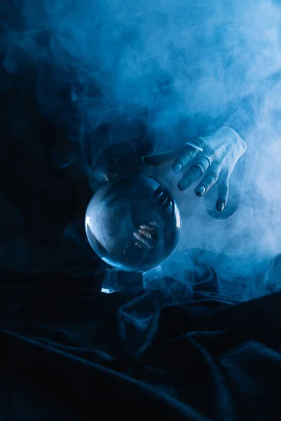 Vue recadrée de la main féminine au-dessus d'une boule de cristal avec fumée sur bleu foncé — Photo de stock