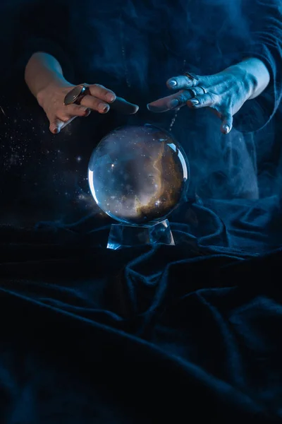 Обрезанный вид ведьмы, выполняющей ритуал с хрустальным шаром на темно-синем — стоковое фото