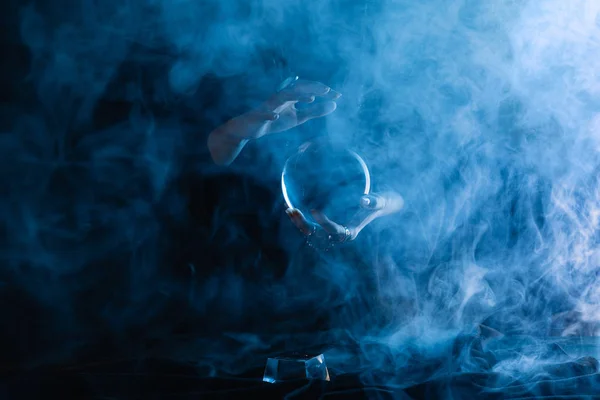 Vista recortada de bruja sosteniendo bola de cristal con humo alrededor en azul oscuro - foto de stock