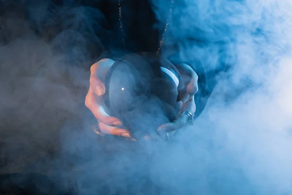 Vista parcial de las manos femeninas con bola de cristal y humo sobre fondo azul oscuro - foto de stock