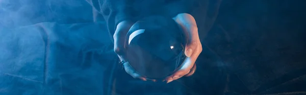 Vista ritagliata della strega che tiene la sfera di cristallo sopra il tavolo su sfondo blu scuro, colpo panoramico — Foto stock