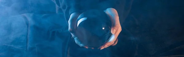 Cropped view of witch holding crystal ball above table on dark blue background, panoramic shot — Stock Photo