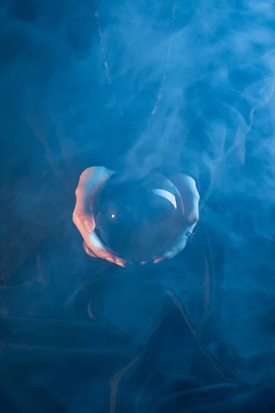 Vista recortada de la bruja sosteniendo la bola de cristal sobre la mesa sobre fondo azul oscuro - foto de stock