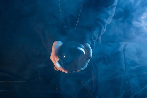 Vue recadrée de la sorcière tenant une boule de cristal au-dessus de la table sur bleu foncé — Photo de stock
