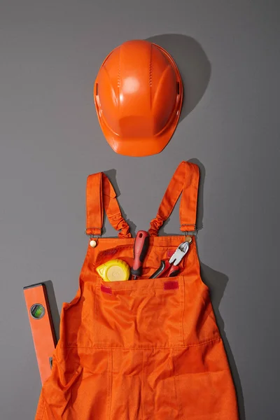 Top view of orange helmet and overalls with measuring tape, screwdriver and pliers near spirit level on grey background — Stock Photo