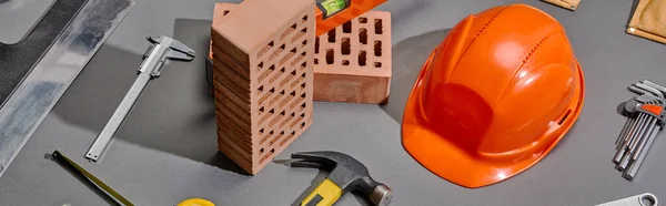 Plano panorámico de ladrillos, martillo, llaves angulares, casco naranja, pinzas y cuchillo de masilla sobre fondo gris - foto de stock
