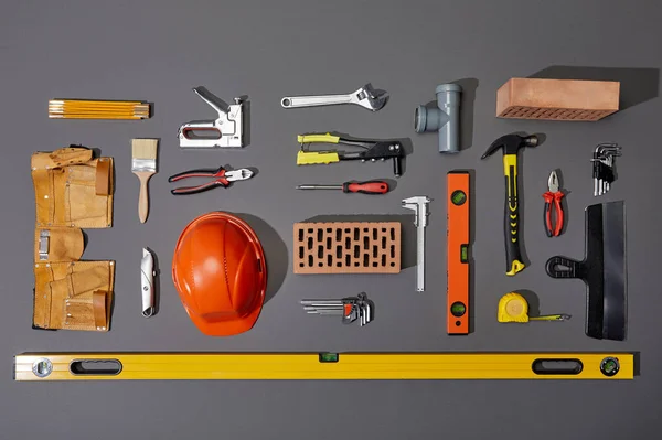 Flat lay with bricks, orange helmet, tool belt and industrial tools on grey background — Stock Photo