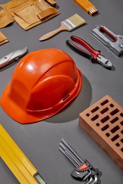 Posa piatta con casco arancione, mattone, pennello, mattone, cintura utensile e strumenti industriali su sfondo grigio — Foto stock