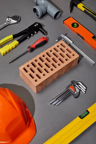 Flat lay with orange helmet, brick, rivet gun, hammer, screwdriver, calipers, spirit level, angle keys and pipe connector on grey background — Stock Photo