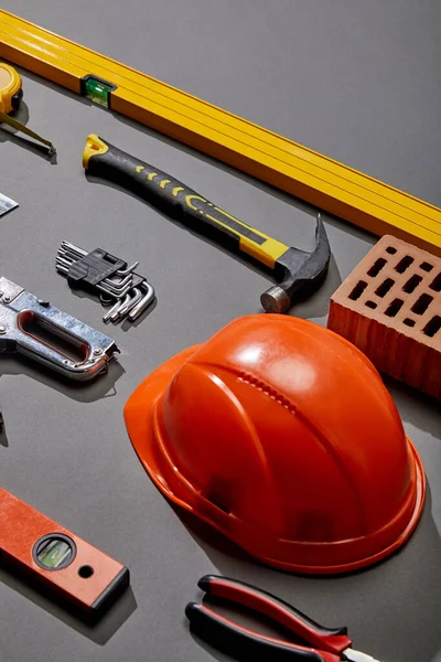 High angle view of orange helmet, hammer, spirit levels, brick, angle keys, pliers, stapler and measuring tape on grey background — Stock Photo