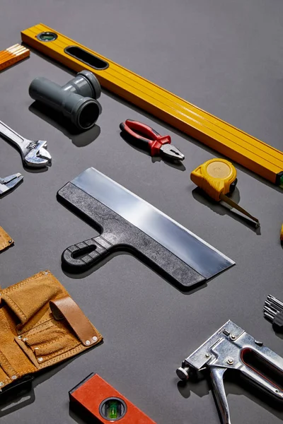 High angle view of putty knife, spirit levels, pipe connector, pliers, measuring tape, monkey wrench, tool belt and stapler on grey background — Stock Photo