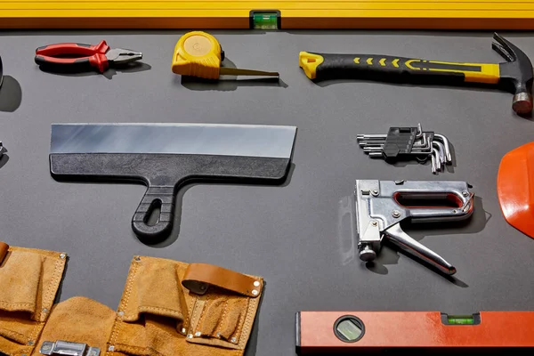 High angle view of spirit levels, pliers, measuring tape, hammer, putty knife, angle keys, stapler and tool belt on grey background — Stock Photo