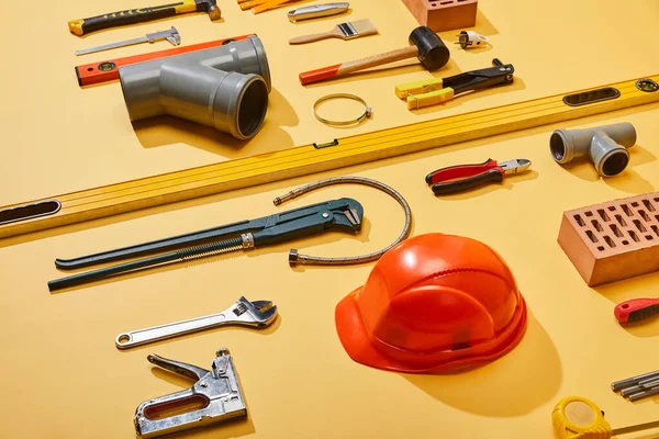 Vista ad alto angolo di utensili industriali, casco, mattoni, nastro di misura, spazzola e tubo idraulico su sfondo giallo — Foto stock