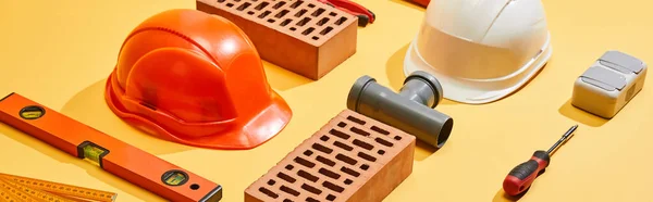 Panoramic shot of bricks, helmets, spirit level, pipe connector, folding ruler, and screwdriver on yellow background — Stock Photo