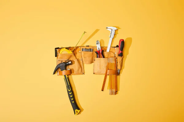 Vista superior do cinto de ferramentas com martelo, alicate, fita métrica, paquímetro, chave de fenda e régua dobrável sobre fundo amarelo — Fotografia de Stock