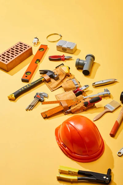 Vista de ángulo alto de la correa de herramientas, herramientas industriales, ladrillo y casco sobre fondo amarillo - foto de stock