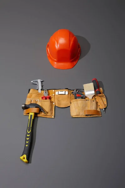 Hochwinkelaufnahme des Helms, Werkzeuggürtels mit Hammer, Pinsel, Zange, Bremssätteln, Winkelschlüsseln und Schraubenzieher auf grauem Hintergrund — Stockfoto