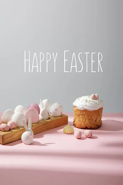 Ostereier auf Holzbrett mit dekorativen Hasen in der Nähe von Osterkuchen auf rosa und grauem Hintergrund mit frohen Osterbildern — Stockfoto