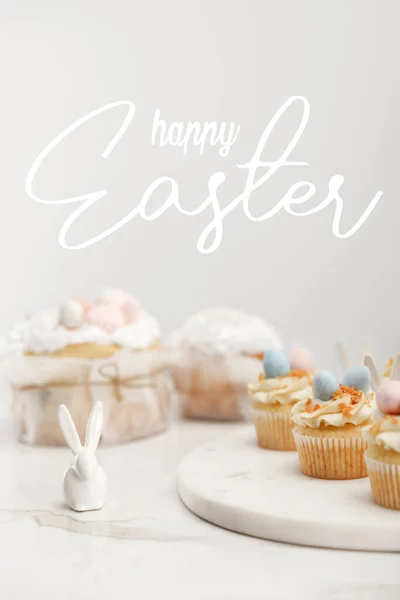 Mise au point sélective de cupcakes sur plateau rond, de lapins décoratifs et de gâteaux de Pâques sur fond gris avec heureuse illustration de Pâques — Photo de stock